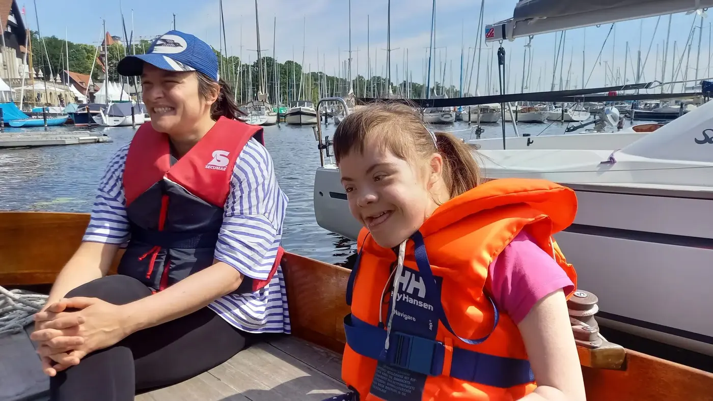Ein Mädchen mit Rettungsweste sitzt strahlend im Cockpit eines Segelboots, neben ihr eine vertraute Betreuerin